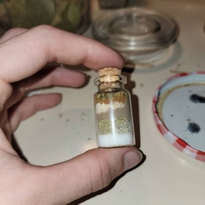 May include: A small, clear glass bottle filled with layered spices and herbs, sealed with a cork stopper. The layers include white, green, and brown ingredients. The bottle is held between fingers.