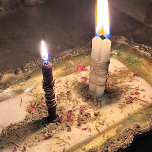 May include: Two lit candles, one purple and one white, are displayed in a decorative tray. The purple candle is wrapped with wire. The white candle is a double wick. The tray contains herbs, rose petals, and a white substance, possibly salt.