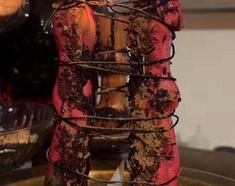 Pode incluir: Duas velas vermelhas cilíndricas e altas estão acesas e posicionadas lado a lado. Estão unidas por um fio fino e escuro e cobertas com uma camada texturizada de ervas e especiarias castanho-escuras. A superfície da cera parece irregular.