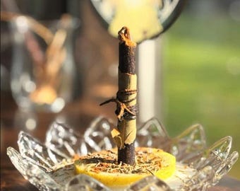 May include: A dark brown candle with a wrapped section, standing on a yellow lemon slice in a clear, decorative glass dish. The candle is adorned with dried herbs and leaves, suggesting a natural or ritualistic theme.