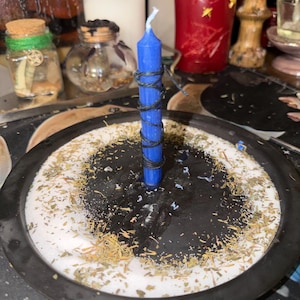 May include: A blue candle wrapped with black twine, centered on a black plate surrounded by white granules and dried herbs. Other candles and small jars are in the background, creating a mystical or spiritual theme.