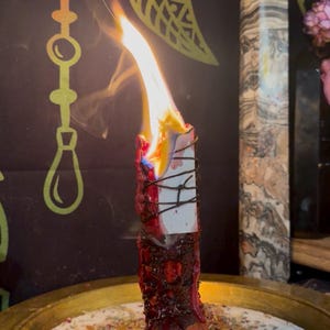 May include: A red pillar candle wrapped in dark twine and burning with a bright flame, positioned in a gold-toned metal tray filled with scattered dried herbs and flower petals, with a pool of melted red wax at the base of the candle.