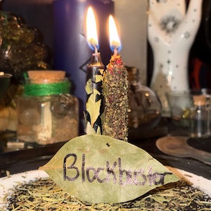 May include: Lit candles with flames, a purple candle, and a leaf with the word "Blockbuster" written on it. The candles are black and brown, with herbs and leaves. The leaf is on a bed of herbs and white powder.