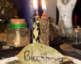 Pode incluir: Velas acesas com chamas, uma vela roxa e uma folha com a palavra "Blockbuster" escrita. As velas são pretas e castanhas, com ervas e folhas. A folha está sobre uma cama de ervas e pó branco.