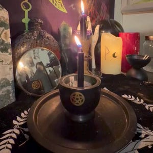 Puede incluir: Una vela negra arde en un cuenco negro con un diseño de pentáculo dorado, sobre una bandeja de metal. En el fondo hay otras velas de varios colores y tamaños, junto con un espejo y cristales. Tema místico.