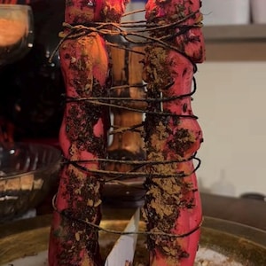 May include: Two tall, cylindrical red candles are burning with bright flames. The candles are wrapped together with thin black string and are covered in a textured layer of brown herbal particles and spices, standing upright on a flat surface.