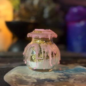 May include: A small, clear glass jar with a rounded body and a cork stopper. The exterior is decorated with pink wax that drips down from the top. Inside the jar, there is a layer of white powder at the bottom topped with dried green herbs.