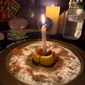 May include: A brass plate holds a white candle centered in a sliced lemon, surrounded by white salt, dried herbs, and reddish-brown powder. In the background, a glass jar labeled Eclipse 006 Moon Water sits near a crystal ball and candle.