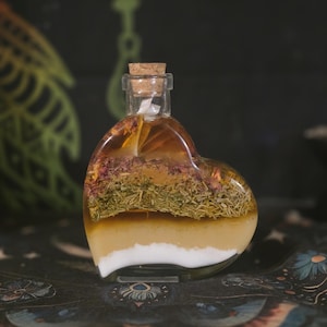 May include: A heart-shaped glass bottle with a cork stopper, filled with layered contents including a white base, a golden liquid, a layer of dried green herbs, and a top layer of dried pink flower petals submerged in a clear amber liquid.