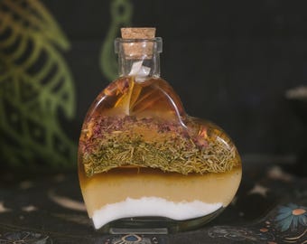 May include: A heart-shaped glass bottle with a cork stopper, filled with layered contents including a white base, a golden liquid, a layer of dried green herbs, and a top layer of dried pink flower petals submerged in a clear amber liquid.