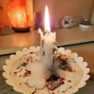 May include: A lit white candle with a unique melted design sits on a scalloped-edge dish. The candle is surrounded by dried herbs and rose petals. The background includes a glowing orange salt lamp and other decorative items.