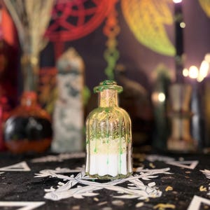 May include: A clear glass bottle with a green-tinted top and dripping green wax. The bottle contains layers of white and light green substances. The background is blurred, featuring other decorative items and a patterned surface.