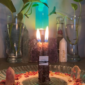 May include: A burning candle with a heart-shaped drawing on a small card, surrounded by crystals and dried rose petals. The candle is on a glass plate, with plants in vases behind it. A lava lamp is also visible.