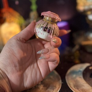 May include: A small, clear glass jar filled with white powder and dark botanical elements. The top of the jar is sealed with pink wax that drips down the sides, creating a textured, layered effect over the glass surface and the contents inside.