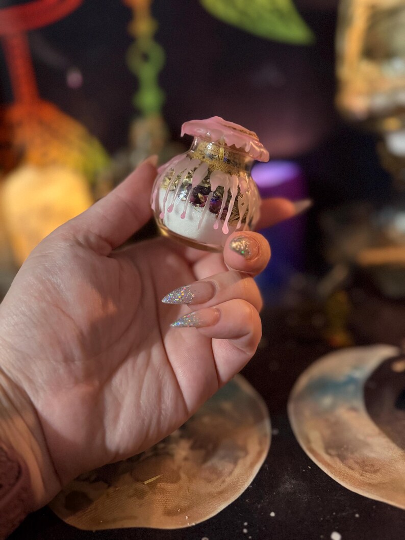 May include: A small, clear glass jar filled with white powder and dark botanical elements. The top of the jar is sealed with pink wax that drips down the sides, creating a textured, layered effect over the glass surface and the contents inside.