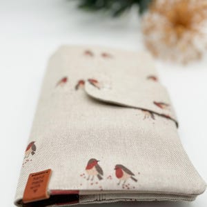 Könnte beinhalten: Eine beige Stoffbörse mit Klappenverschluss. Die Börse hat ein Muster aus roten Rotkehlchen auf weißem Hintergrund. Die Börse hat einen braunen Lederlasche mit dem Text "Made with Love" darauf.