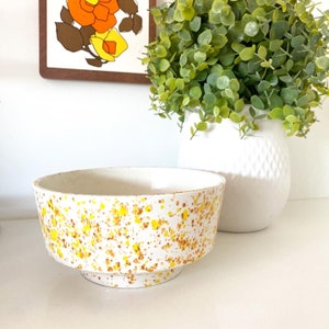May include: A white ceramic bowl with a yellow and orange speckled design. The bowl has a wide base and a rounded top.