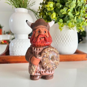 May include: A ceramic figurine of a Viking warrior with a red beard and a shield. The figurine is painted in brown, red, and white.