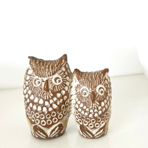 May include: Two ceramic owls, one larger than the other, are painted in brown and white with a textured surface. The owls are facing forward with their wings touching.