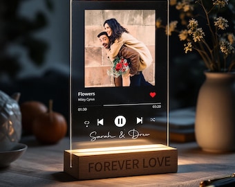 May include: A clear acrylic photo frame with a wooden base. The frame has a picture of a couple in the background and a music player interface with the song "Flowers" by Miley Cyrus playing. The base has the text "Forever Love" engraved on it.