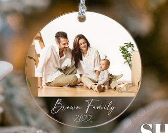 May include: Round, clear, glass Christmas ornament with a photo of a family of three. The photo shows a man, a woman, and a baby. The ornament has a white string for hanging and the text "Brown Family 2022" printed on the bottom.
