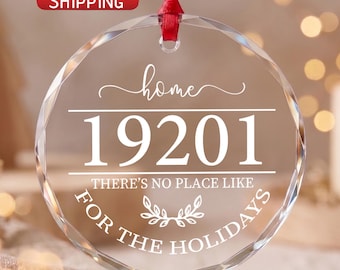 May include: A round, faceted clear glass ornament hanging from a red ribbon. The ornament features white text that reads home, 19201, and THERE'S NO PLACE LIKE FOR THE HOLIDAYS, with a small leaf graphic centered between the lines of text.