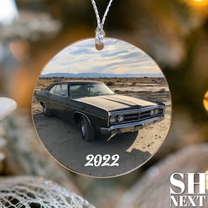May include: A round, clear, glass ornament with a photo of a dark green 1960s era car in a desert setting. The year 2022 is printed in white on the ornament.