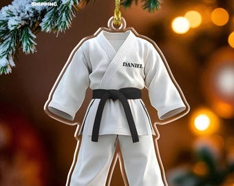 May include: A white martial arts uniform ornament featuring a black belt and the name DANIEL printed in black on the chest. The flat, shaped acrylic decoration hangs from a gold string, designed to resemble a traditional karate gi outfit.