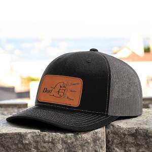 May include: Black and gray trucker hat with a brown leather patch that says "Dad" and has four names listed below it: Cameron, Hunter, Reagan.