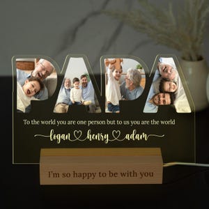 May include: A personalized illuminated acrylic sign with the word "PAPA" in large letters. The sign features multiple photos of a man with his children and grandchildren. The text "To the world you are one person but to us you are the world" is written below the photos, followed by the names "logan henry adam" with heart symbols between each name. The sign also includes the text "I'm so happy to be with you" on a wooden base.