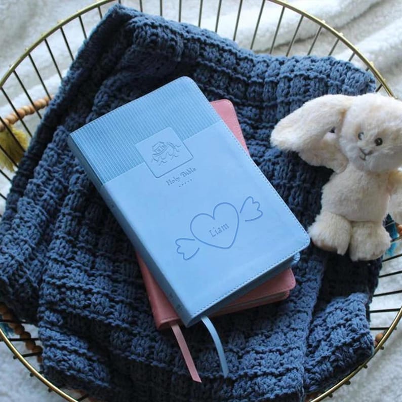 Pu&ograve; includere: Una Bibbia per bambini di colore azzurro chiaro con un cuore bianco e il nome "Liam" sulla copertina. La Bibbia &egrave; appoggiata su una coperta di maglia blu con un coniglietto di peluche bianco.
