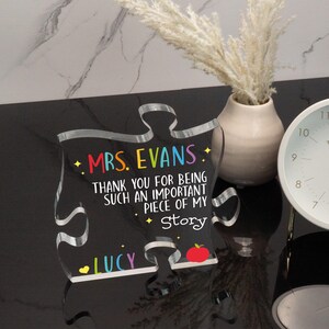 K&ouml;nnte beinhalten: Ein klares Acryl-Puzzleteil mit schwarzem Hintergrund und buntem Text, der "MRS. EVANS. THANK YOU FOR BEING SUCH AN IMPORTANT PIECE OF MY Story LUCY" lautet. Am unteren Rand des Teils befindet sich eine rote Apfelgrafik.