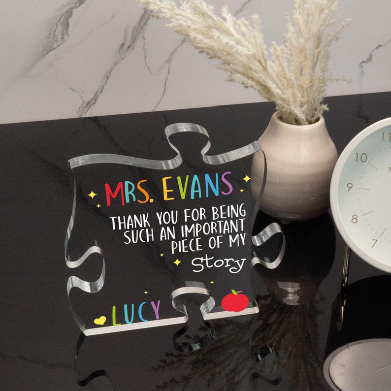 K&ouml;nnte beinhalten: Ein klares Acryl-Puzzleteil mit schwarzem Hintergrund und buntem Text, der "MRS. EVANS. THANK YOU FOR BEING SUCH AN IMPORTANT PIECE OF MY Story LUCY" lautet. Am unteren Rand des Teils befindet sich eine rote Apfelgrafik.