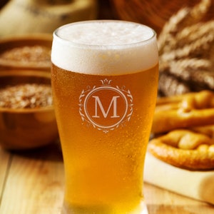 May include: A frosted glass beer mug with a monogrammed "M" in a decorative frame. The mug is filled with a golden-colored beer and has a white head. The mug is sitting on a wooden table.