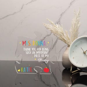 K&ouml;nnte beinhalten: Ein klares Acryl-Puzzleteil mit dem Text "MRS. DONNA" in Regenbogenfarben. Der Text "THANK YOU FOR BEING SUCH AN IMPORTANT PIECE OF MY STORY" befindet sich unter dem Namen. Der Name "KATIE" befindet sich unten auf dem Teil mit einem roten Apfel.