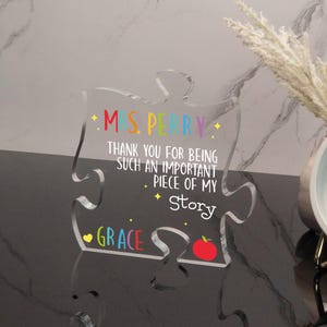 K&ouml;nnte beinhalten: Ein klares Acryl-Puzzleteil mit dem Text "MS. PERRY" in Regenbogenfarben. Der Text "THANK YOU FOR BEING SUCH AN IMPORTANT PIECE OF MY STORY" ist ebenfalls enthalten. Am unteren Rand des Teils steht der Name "GRACE" und ein roter Apfel.