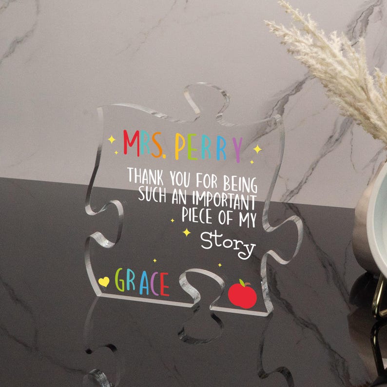 K&ouml;nnte beinhalten: Ein klares Acryl-Puzzleteil mit dem Text "MS. PERRY" in Regenbogenfarben. Der Text "THANK YOU FOR BEING SUCH AN IMPORTANT PIECE OF MY STORY" ist ebenfalls enthalten. Am unteren Rand des Teils steht der Name "GRACE" und ein roter Apfel.