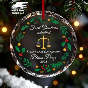 May include: A circular glass Christmas ornament with a faceted edge, featuring a gold scale of justice icon, a green and red holly wreath design, and white text reading First Christmas admitted, State Bar of Georgetown, Brian Perez, 2025.