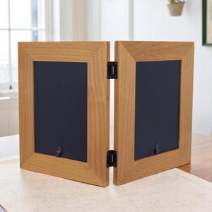 May include: A two-panel wooden picture frame with a light brown finish. The frame is connected by black hinges and has a dark blue interior. The frame is designed to stand upright on a table or shelf.
