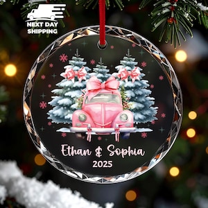 May include: A round, clear glass Christmas ornament with a pink car and Christmas trees design. The ornament has the names "Ethan & Sophia" and the year "2025" printed on it. A red ribbon is attached for hanging.