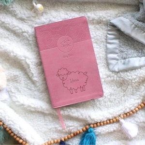 Pu&ograve; includere: Una Bibbia per bambini rosa con un'illustrazione di un agnello e il nome "Maria" in rilievo sulla copertina.
