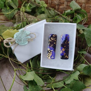 May include: A pair of rectangular resin earrings with a blue and black marbled pattern and gold flecks. The earrings are in a white box.