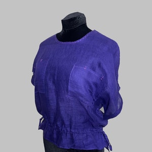 Puede incluir: Una blusa de lino morada con mangas cortas y cintura con cordón. La blusa tiene dos bolsillos en la parte delantera y costuras rosas alrededor del cuello.