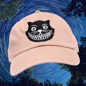 May include: A light pink baseball cap featuring a black Cheshire Cat design. The cat's face is embroidered on the front, with a wide grin and large eyes. The cap has a curved brim and an adjustable strap.