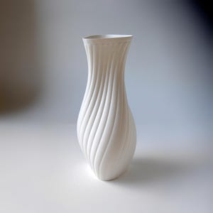 Puede incluir: Un jarr&oacute;n blanco alto que presenta una textura acanalada en espiral distintiva que recorre verticalmente todo su cuerpo. El recipiente tiene un cuello estrecho que se ensancha ligeramente en la abertura superior, creando una silueta elegante.