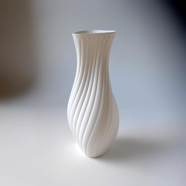 Puede incluir: Un jarr&oacute;n blanco alto que presenta una textura acanalada en espiral distintiva que recorre verticalmente todo su cuerpo. El recipiente tiene un cuello estrecho que se ensancha ligeramente en la abertura superior, creando una silueta elegante.