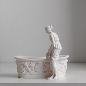 May include: A white matte finish decorative vessel shaped like an oval bathtub with ornate floral and leaf relief carvings on the exterior. A sculpted figure stands beside the tub, one leg positioned inside, wearing a draped garment.