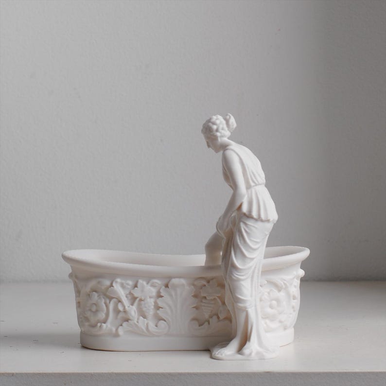 May include: A white matte finish decorative vessel shaped like an oval bathtub with ornate floral and leaf relief carvings on the exterior. A sculpted figure stands beside the tub, one leg positioned inside, wearing a draped garment.