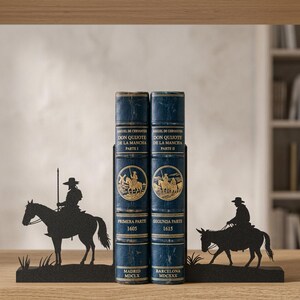 Puede incluir: Dos libros de tapa dura azul oscuro titulados Don Quijote de la Mancha, Parte I y Parte II, est&aacute;n sujetos por dos sujetalibros de metal negro. El sujetalibros izquierdo muestra la silueta de un jinete a caballo, y el derecho un jinete sobre un burro.