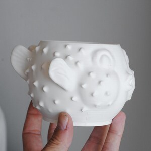 Puede incluir: Una taza de cer&aacute;mica blanca mate con forma de pez globo, que presenta un cuerpo redondeado con espinas en relieve, peque&ntilde;as aletas que sobresalen por los lados y un detalle de boca fruncida en la parte frontal. La superficie es lisa y uniforme.